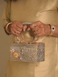 The silver filigree box purse held by its oval handle, charm studded lid facing forward.