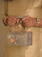 The silver filigree box purse held by its oval handle, charm studded lid facing forward.