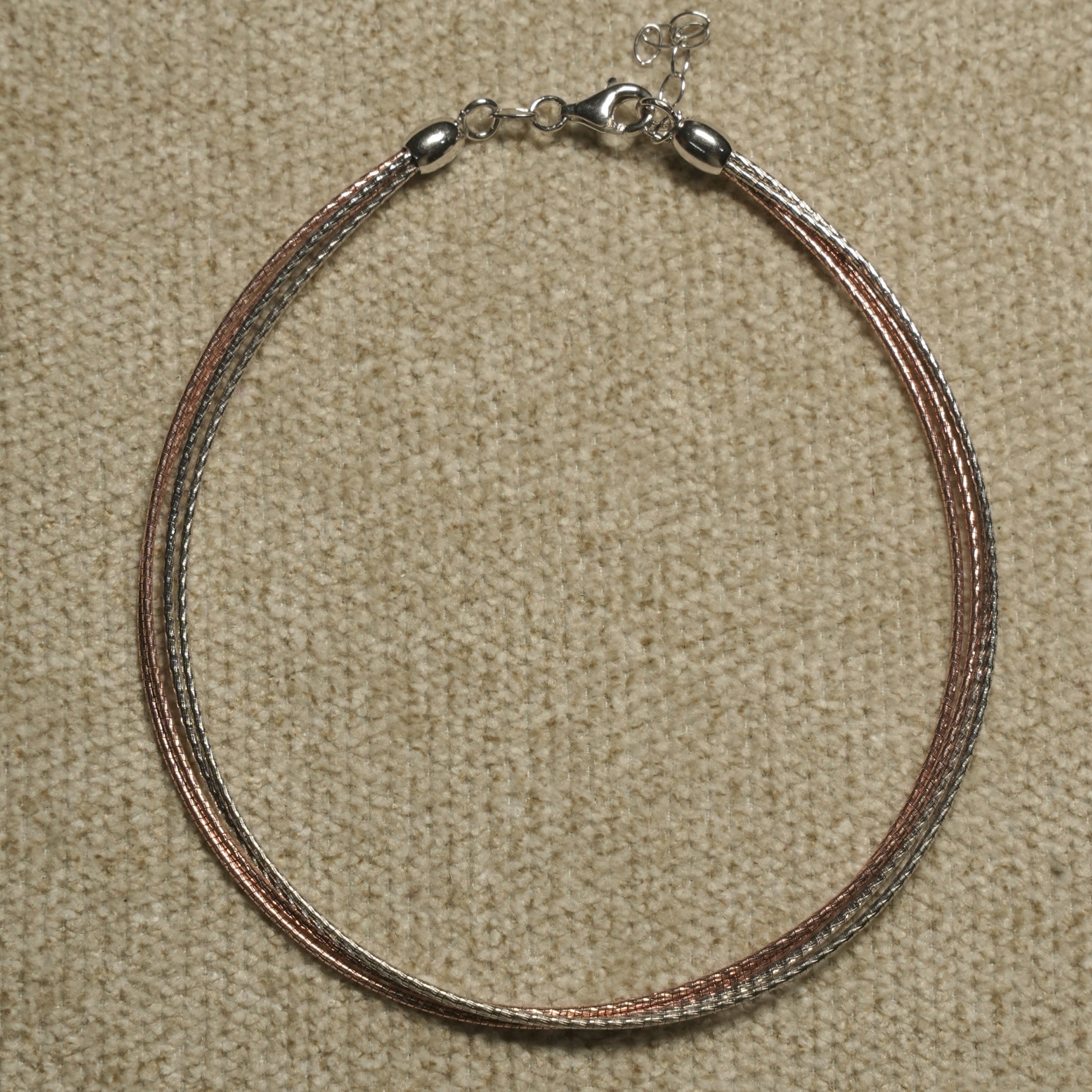 Multi-strand anklet of fine textured silver and rose gold-tone strands with a lobster clasp and extender chain, laid flat on beige fabric.