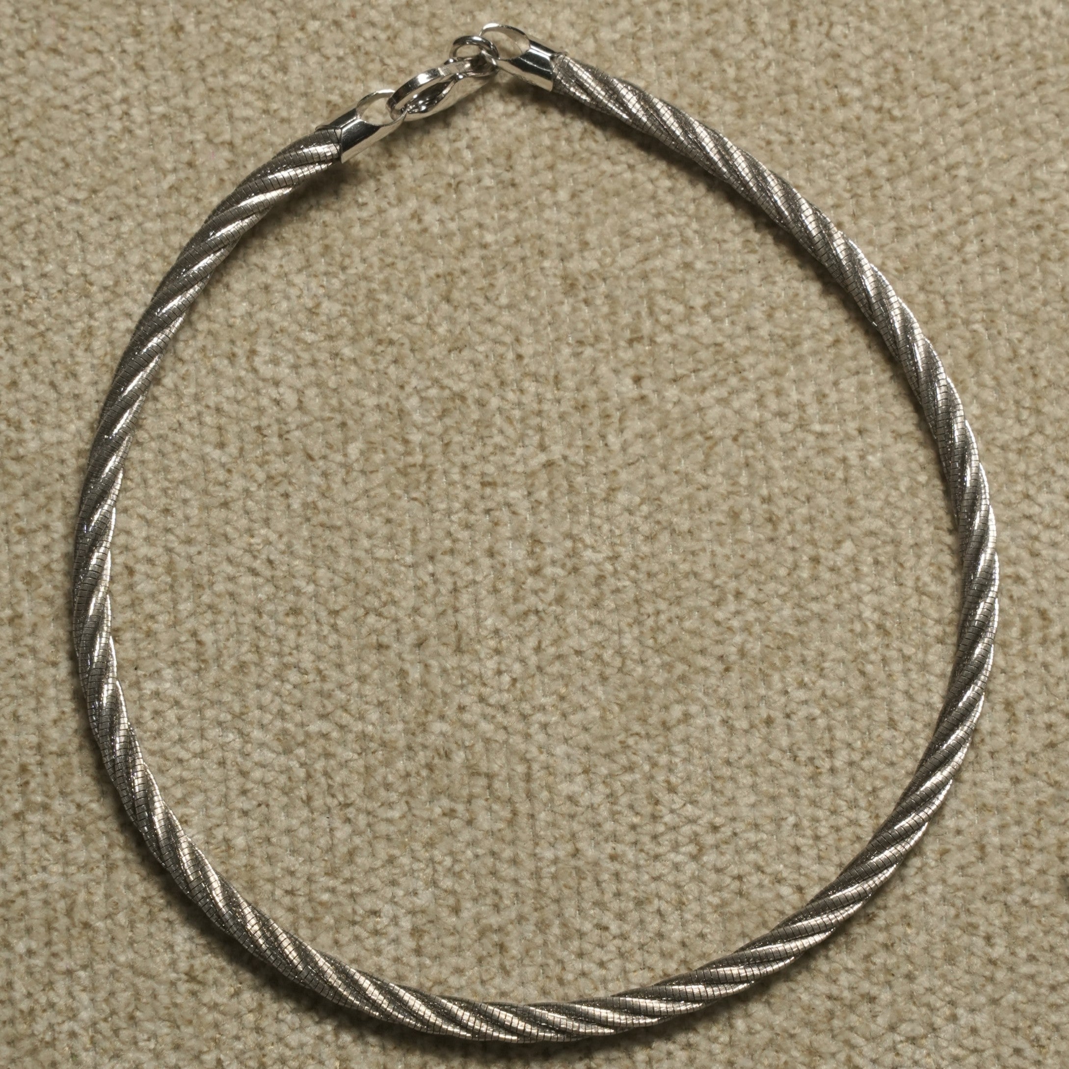 Sterling silver anklet with a twisted rope texture and slender clasp, photographed on beige woven fabric.