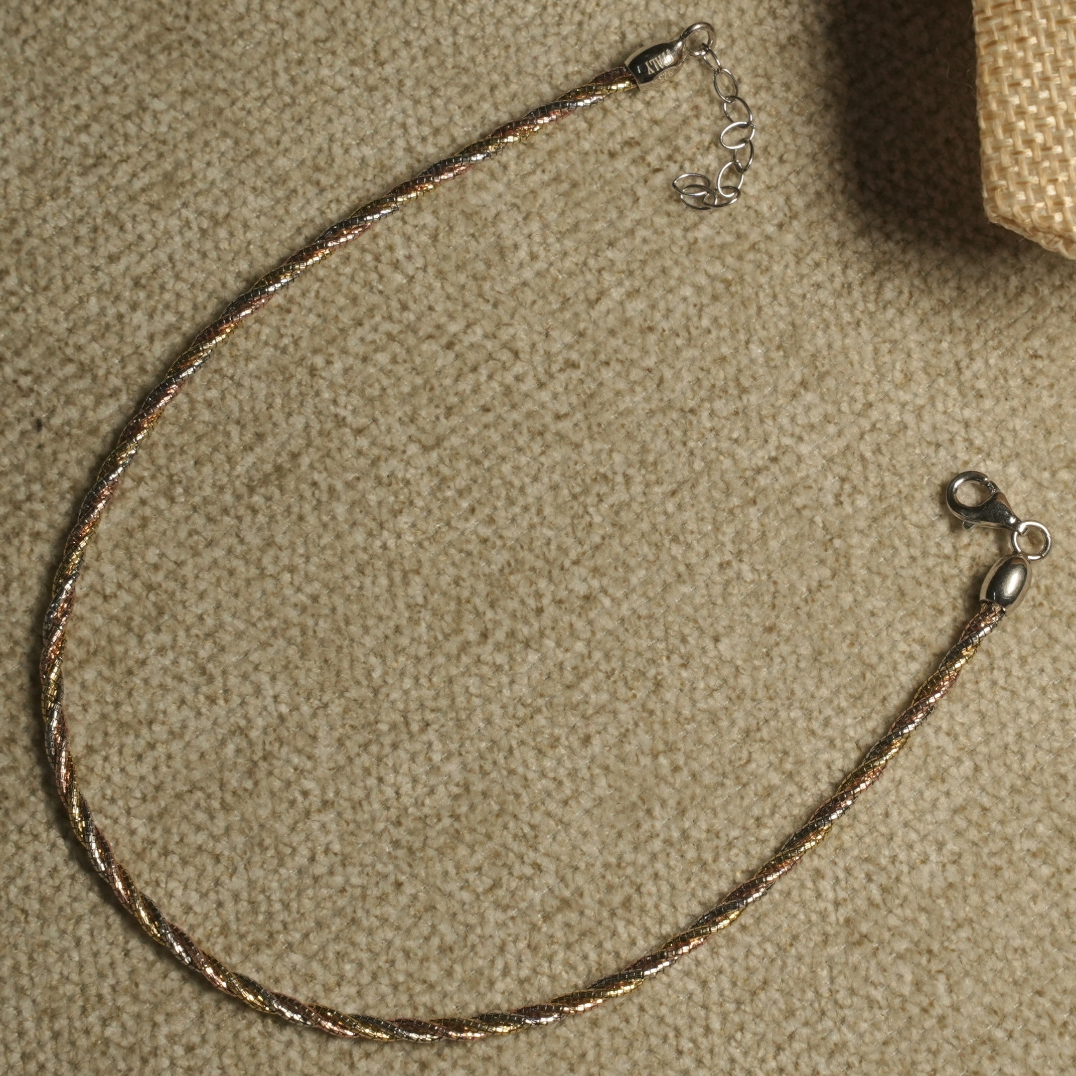 Twisted tri-tone sterling silver anklet with lobster clasp and extender chain laid in a loop on beige fabric.