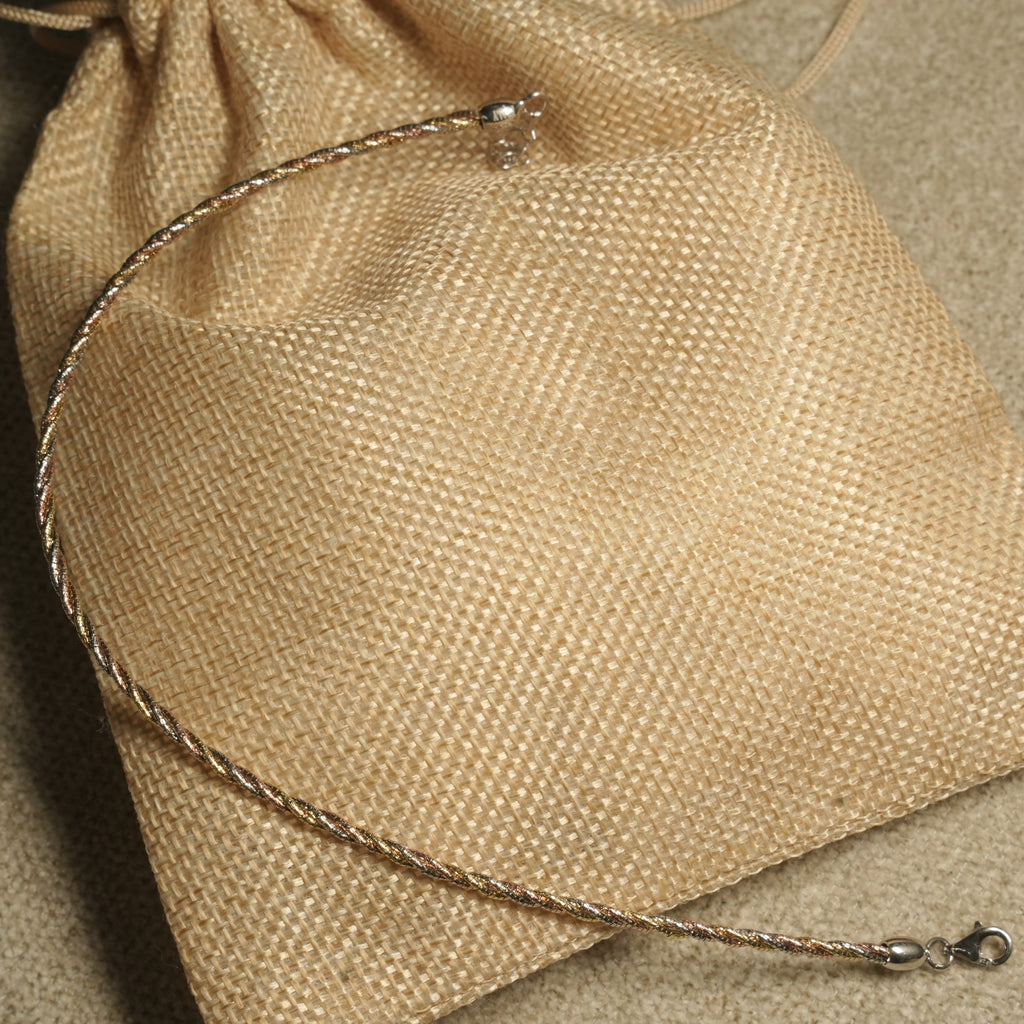 Twisted rope-texture anklet in silver, gold-tone and rose gold-tone resting across a jute pouch.