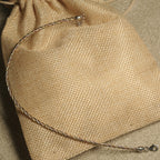Twisted rope-texture anklet in silver, gold-tone and rose gold-tone resting across a jute pouch.