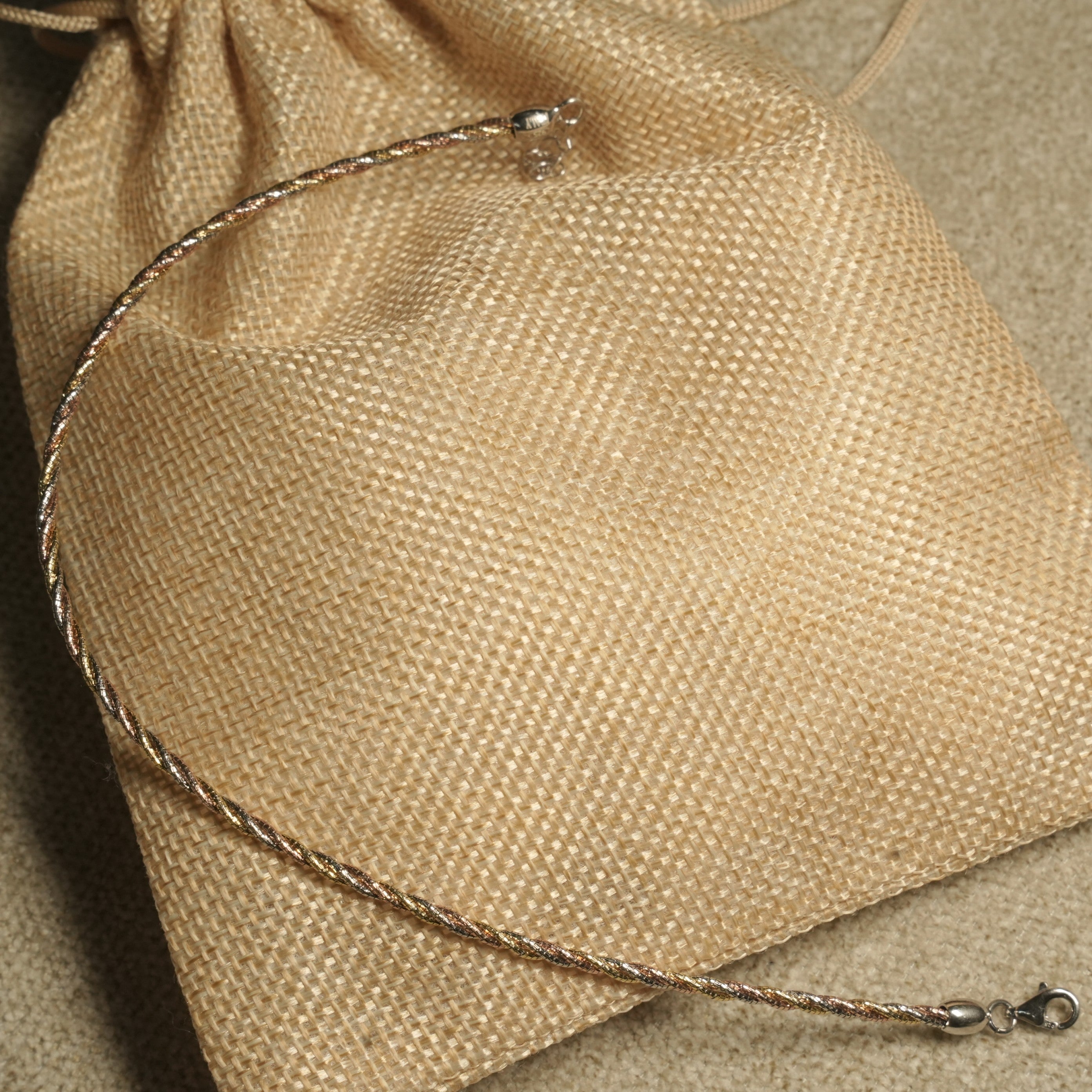 Twisted rope-texture anklet in silver, gold-tone and rose gold-tone resting across a jute pouch.