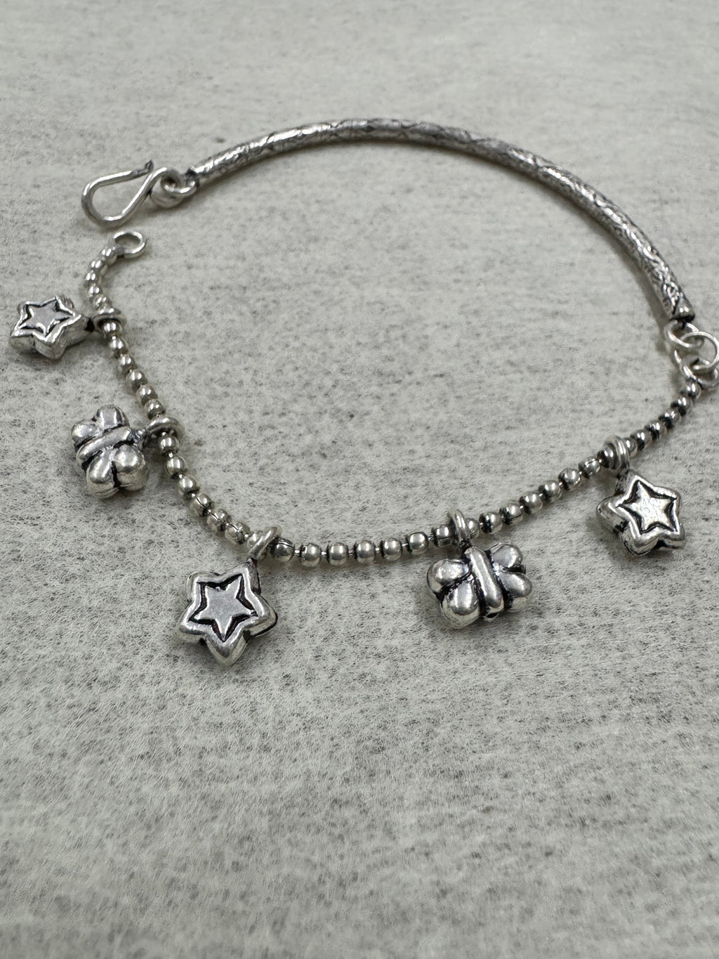 Sterling silver anklet on grey felt, with star and butterfly charms hanging from the ball chain and the engraved bar curving away at the left.