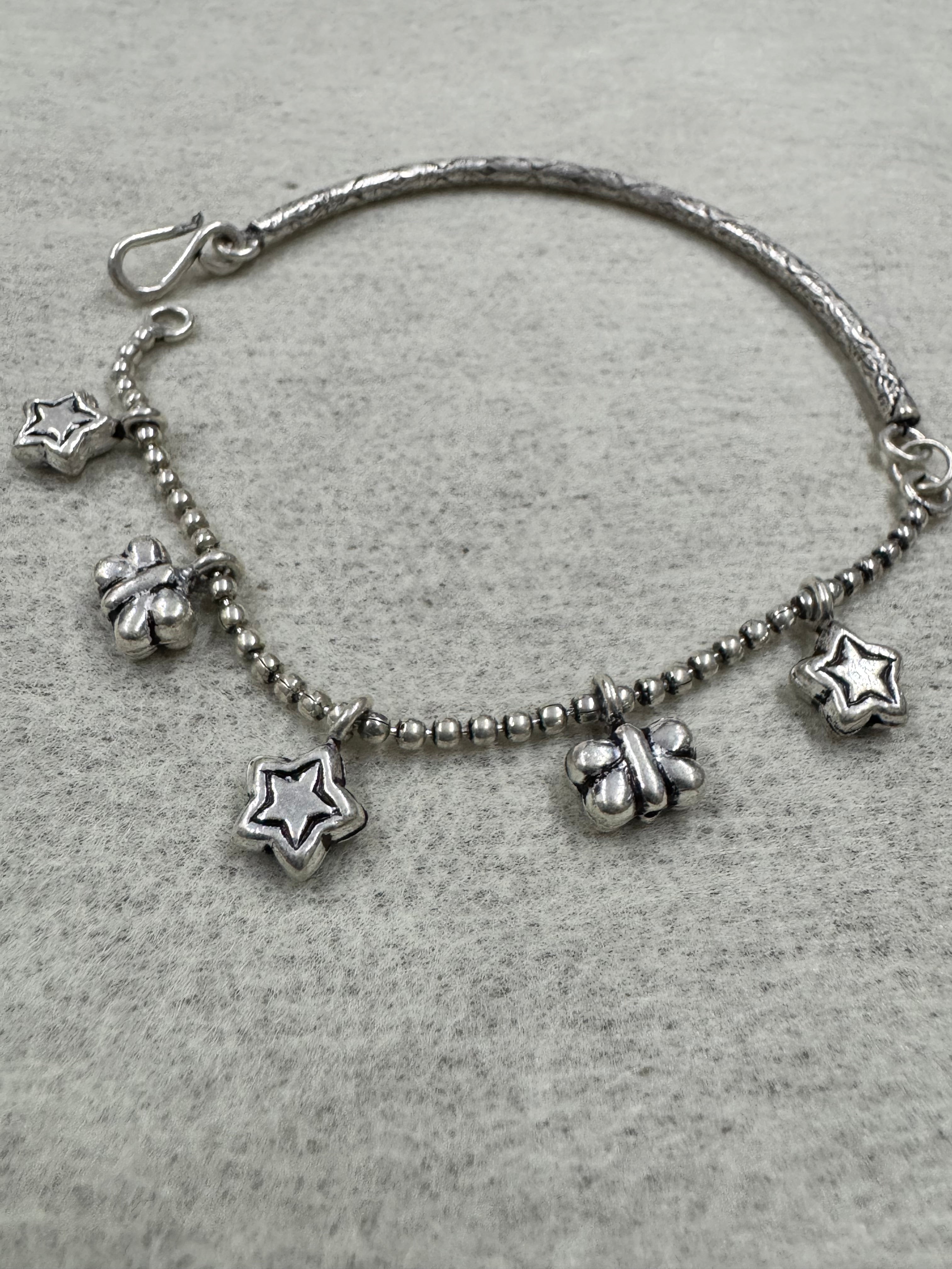 Sterling silver anklet on grey felt, with star and butterfly charms hanging from the ball chain and the engraved bar curving away at the left.