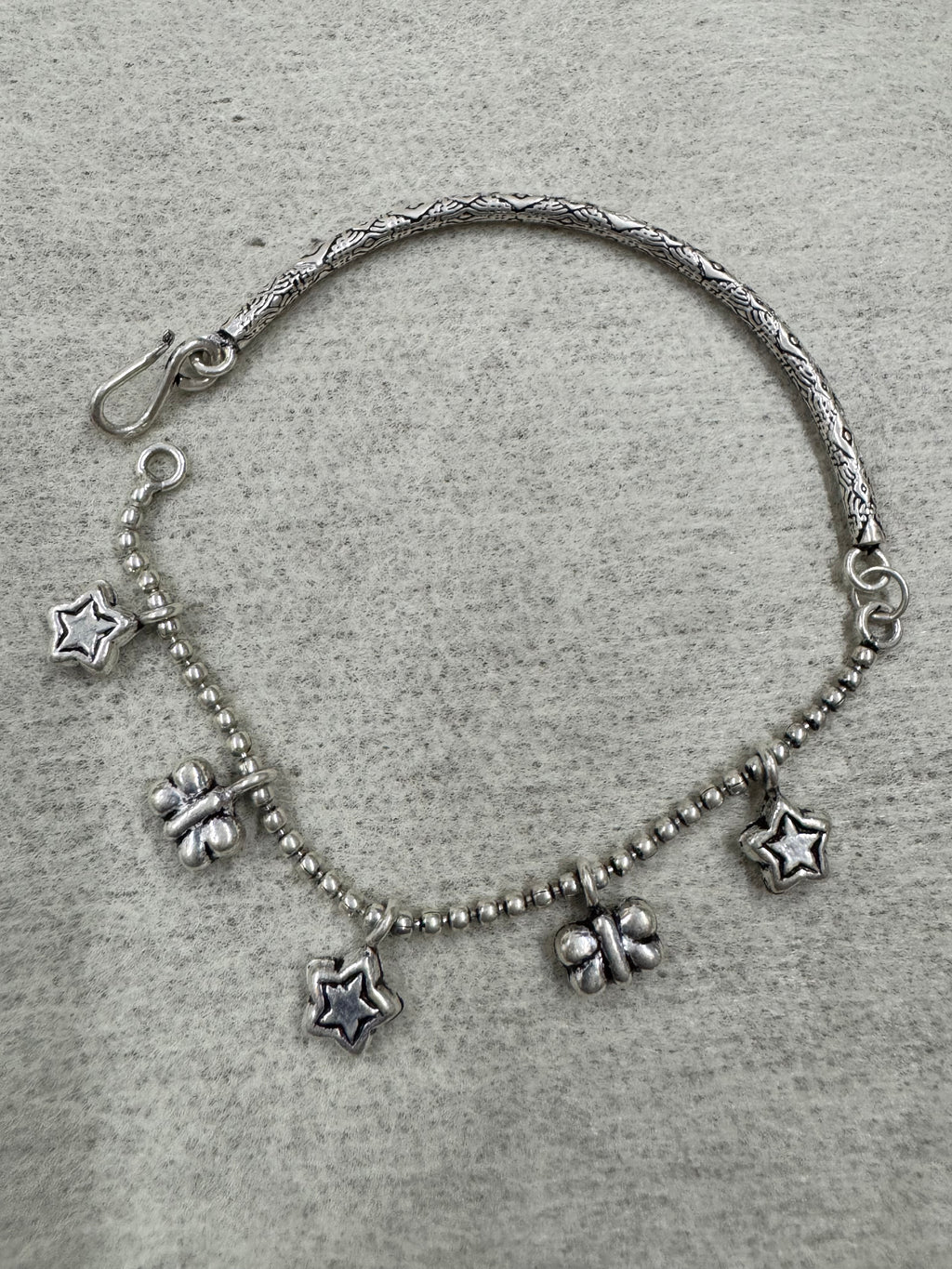 Full flat-lay of the sterling silver charm anklet on grey felt, engraved bar on one side and ball chain with five charms on the other.