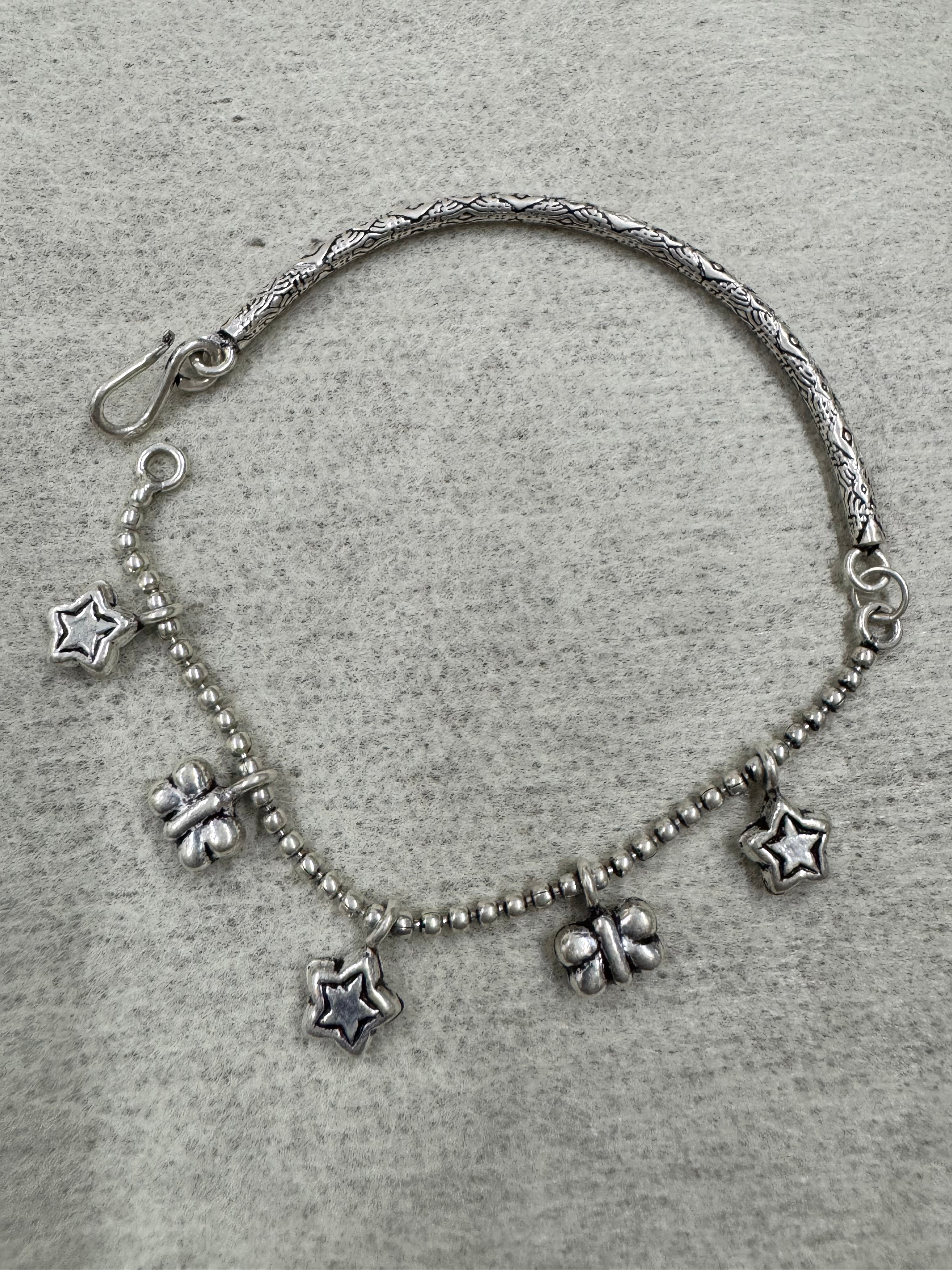 Full flat-lay of the sterling silver charm anklet on grey felt, engraved bar on one side and ball chain with five charms on the other.