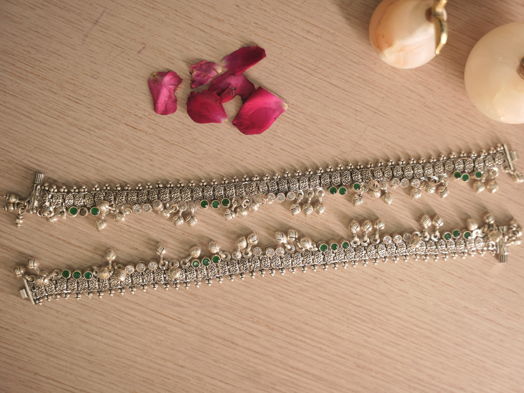 Pair of broad oxidised silver payal with leaf shaped drops and ghungroos, laid out on a pale surface.