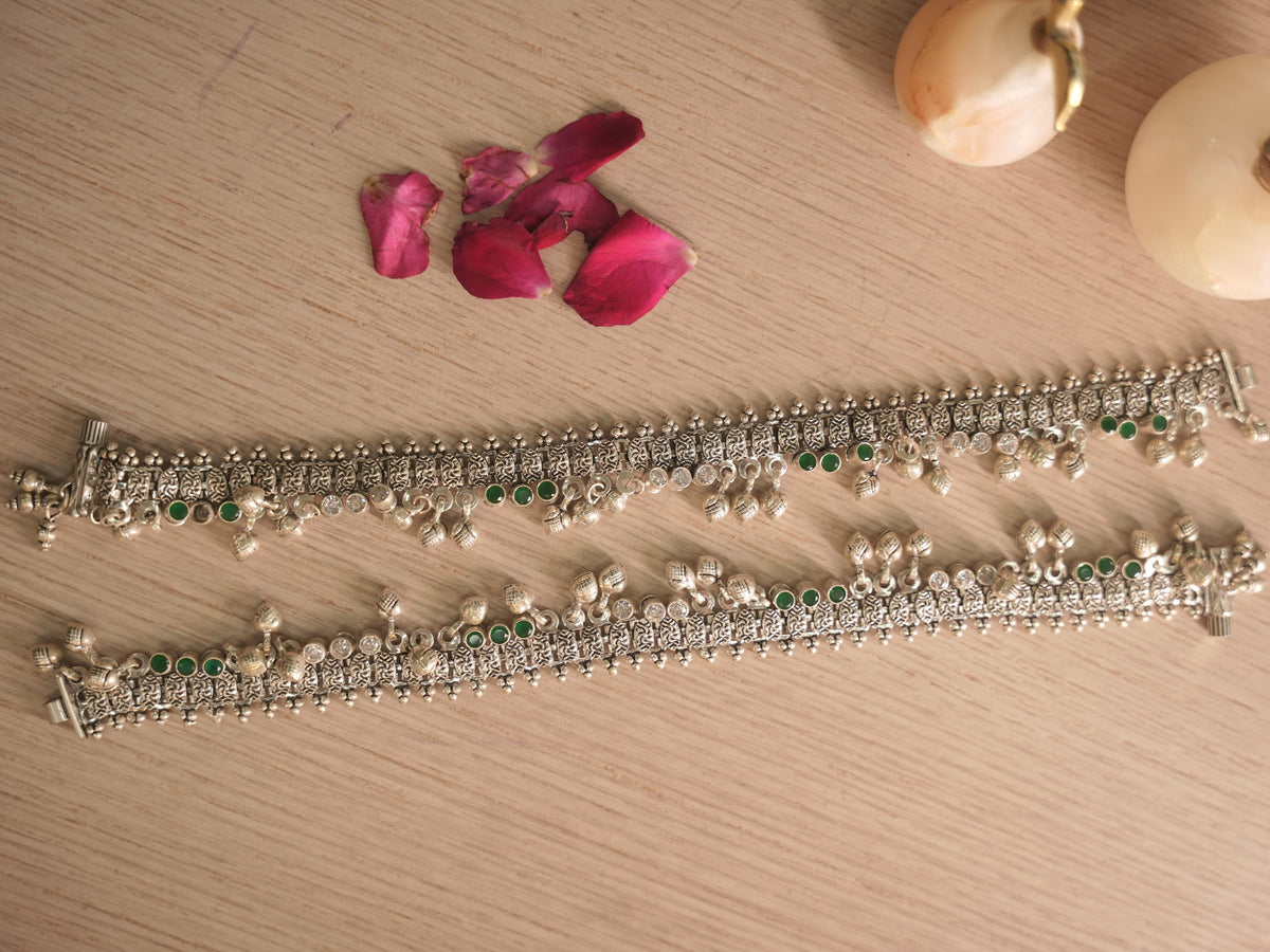 Pair of broad oxidised silver payal with leaf shaped drops and ghungroos, laid out on a pale surface.