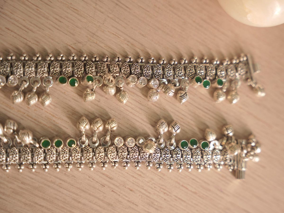 Both oxidised silver payal overlapping, showing the woven band and the spacing of green and colourless stones.