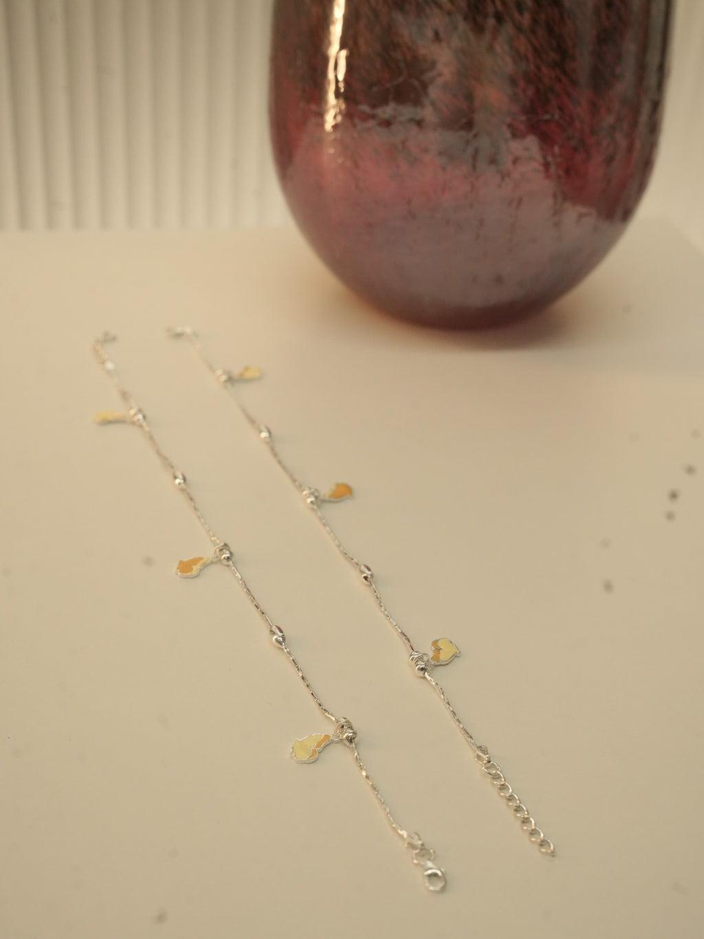 The pair of butterfly payal laid out beside a dark red glass vase.
