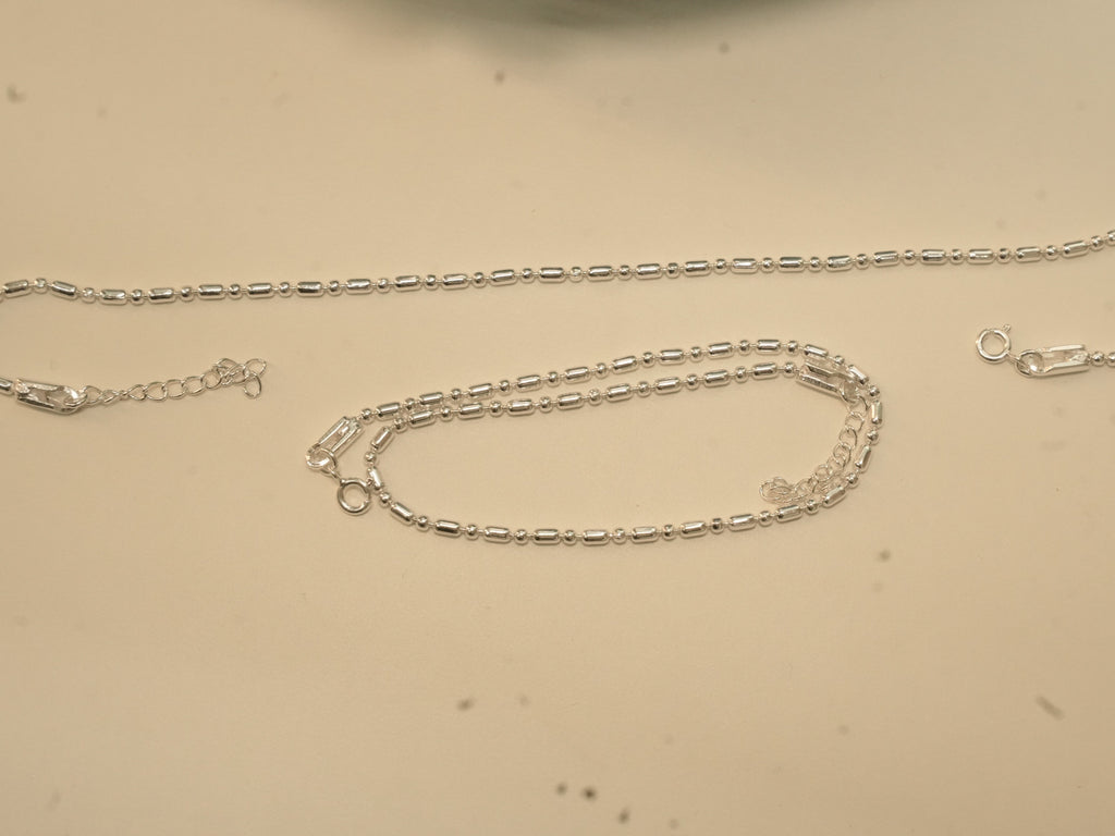 Pair of plain silver payal in a bar and bead chain, one laid straight and one coiled.