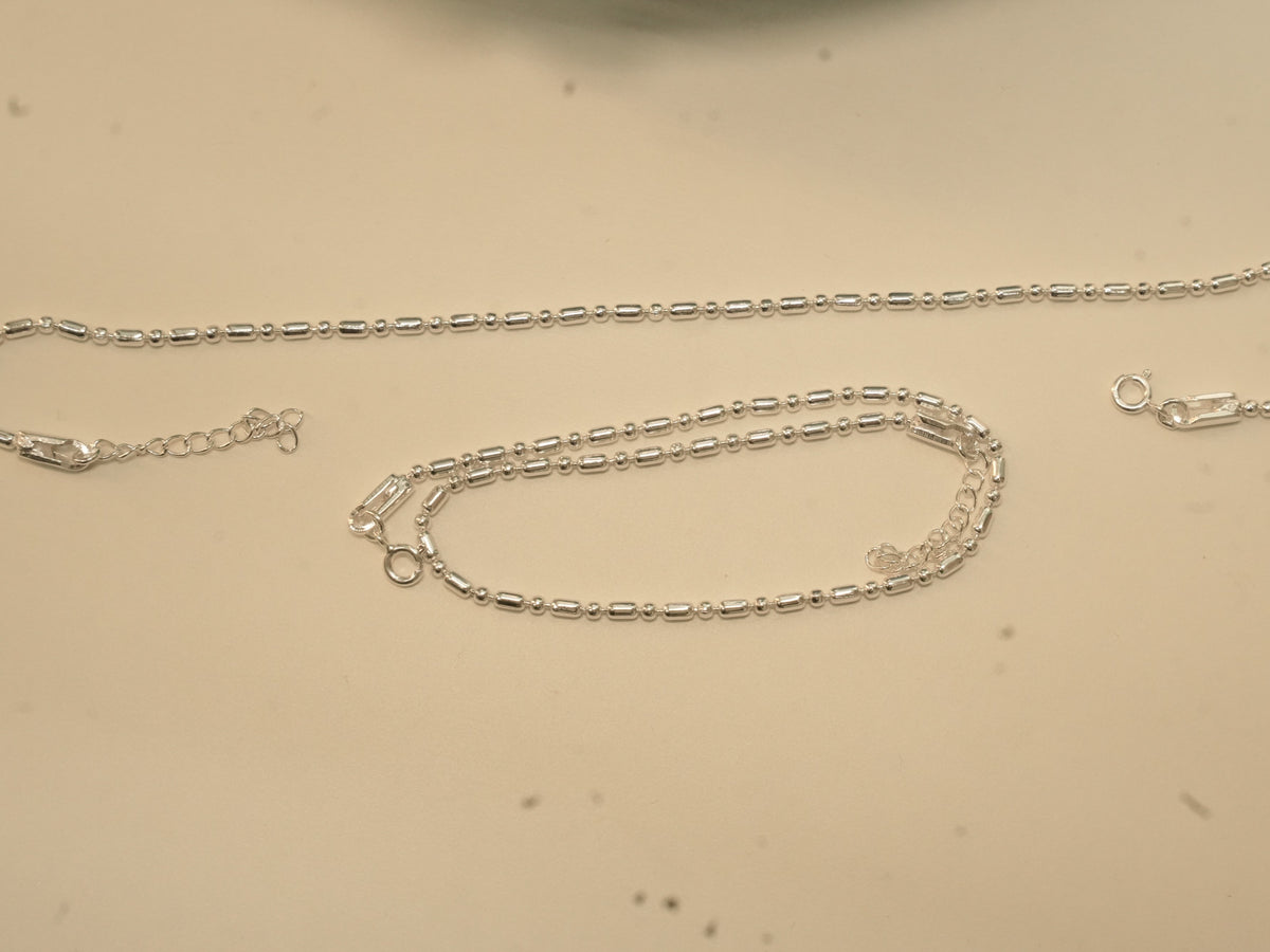 Pair of plain silver payal in a bar and bead chain, one laid straight and one coiled.