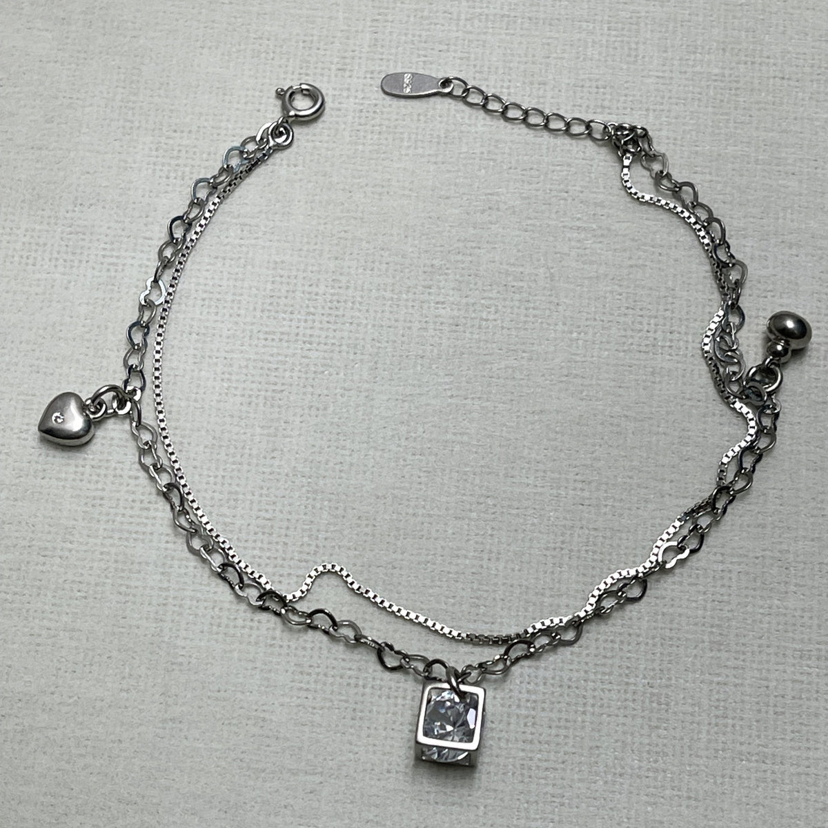 Sterling silver double-strand anklet with a heart charm, a polished ball and an American Diamond inside an open cube, on a pale surface.