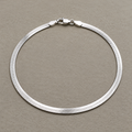 Sterling silver flat herringbone chain anklet laid in a loop with its lobster clasp, on a beige surface.