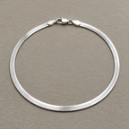 Sterling silver flat herringbone chain anklet laid in a loop with its lobster clasp, on a beige surface.