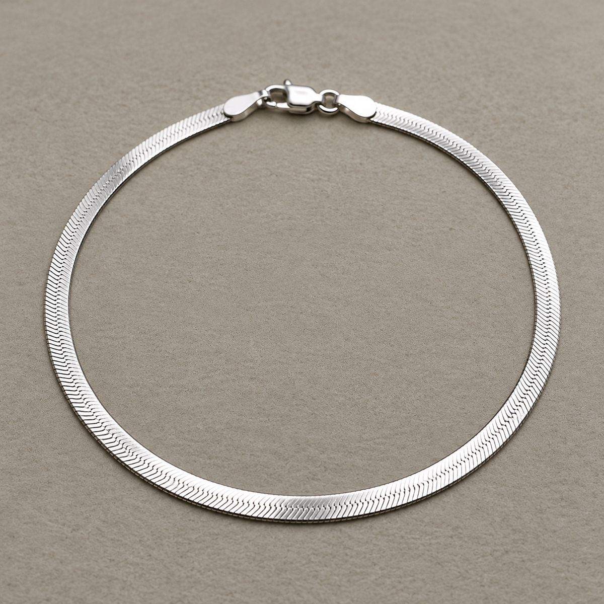 Sterling silver flat herringbone chain anklet laid in a loop with its lobster clasp, on a beige surface.