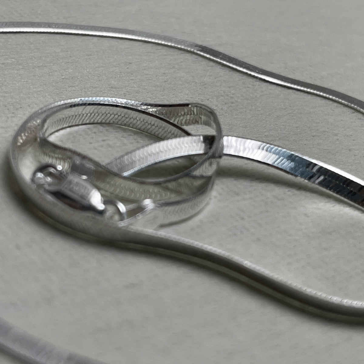 The flat herringbone silver anklet coiled to show the smooth ribbon of links.