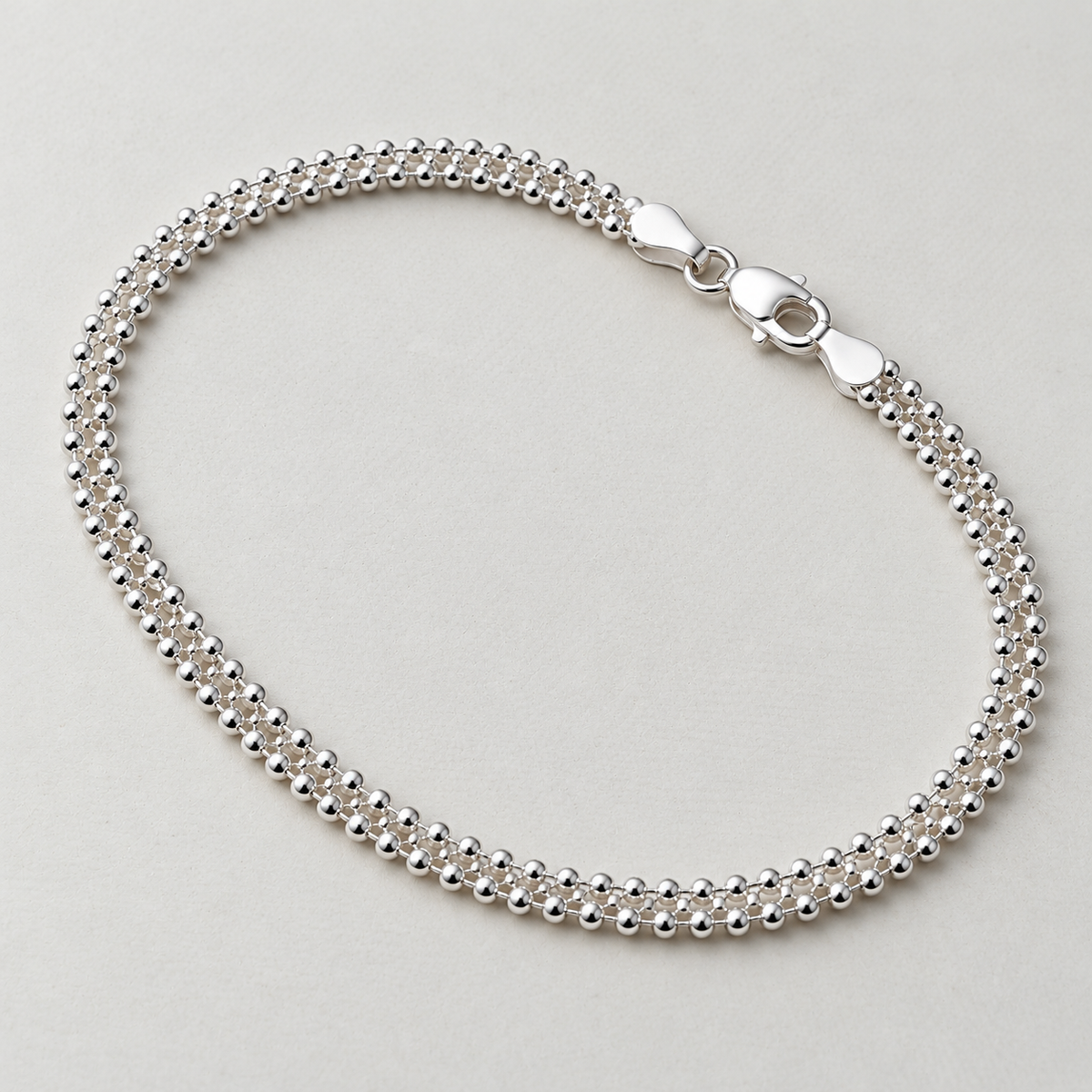 Sterling silver popcorn chain anklet laid in a loop with its lobster clasp, on a cream surface.