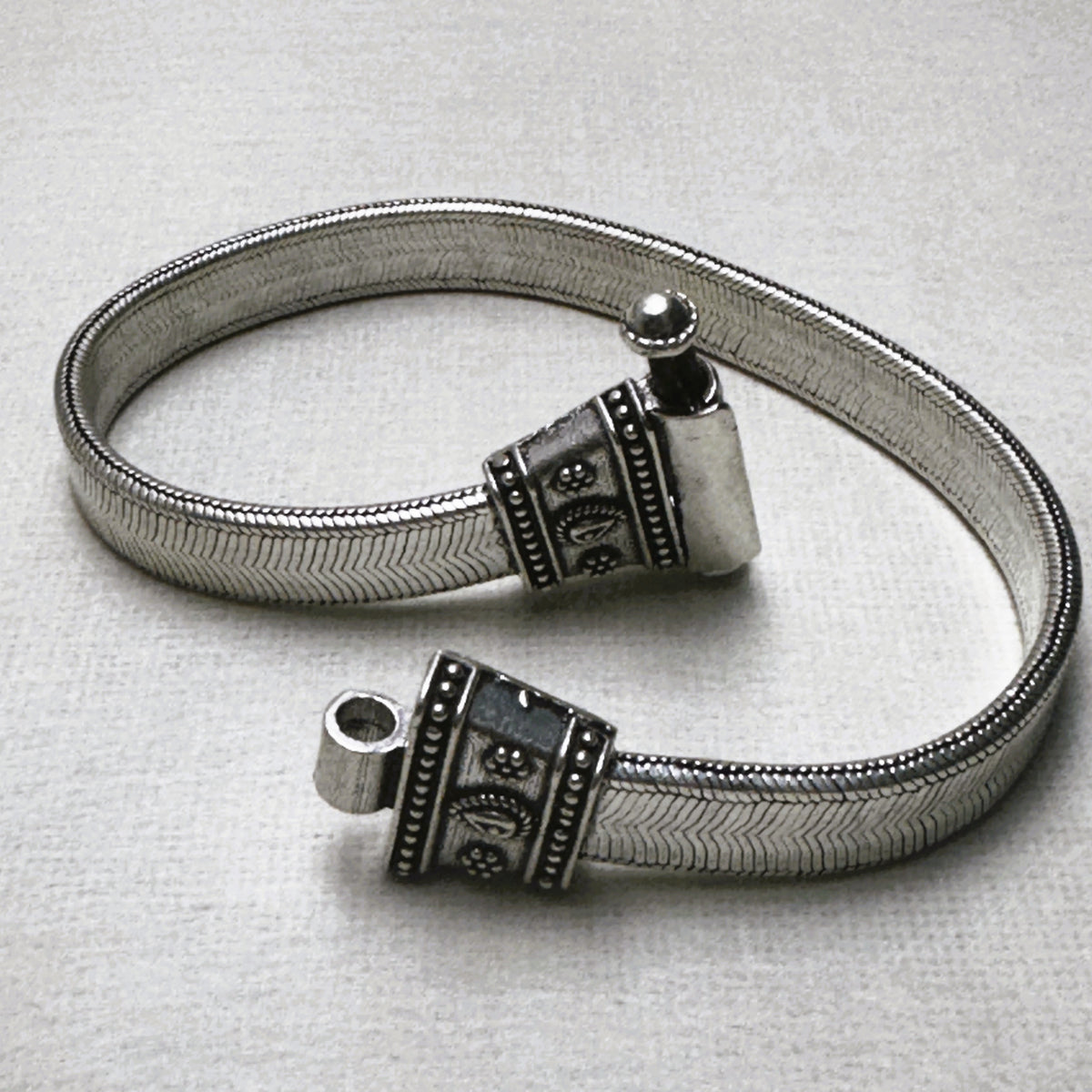 Rajwadi sterling silver payal, a wide flat snake chain with carved oxidised end caps, laid in a loop with the pin clasp open.
