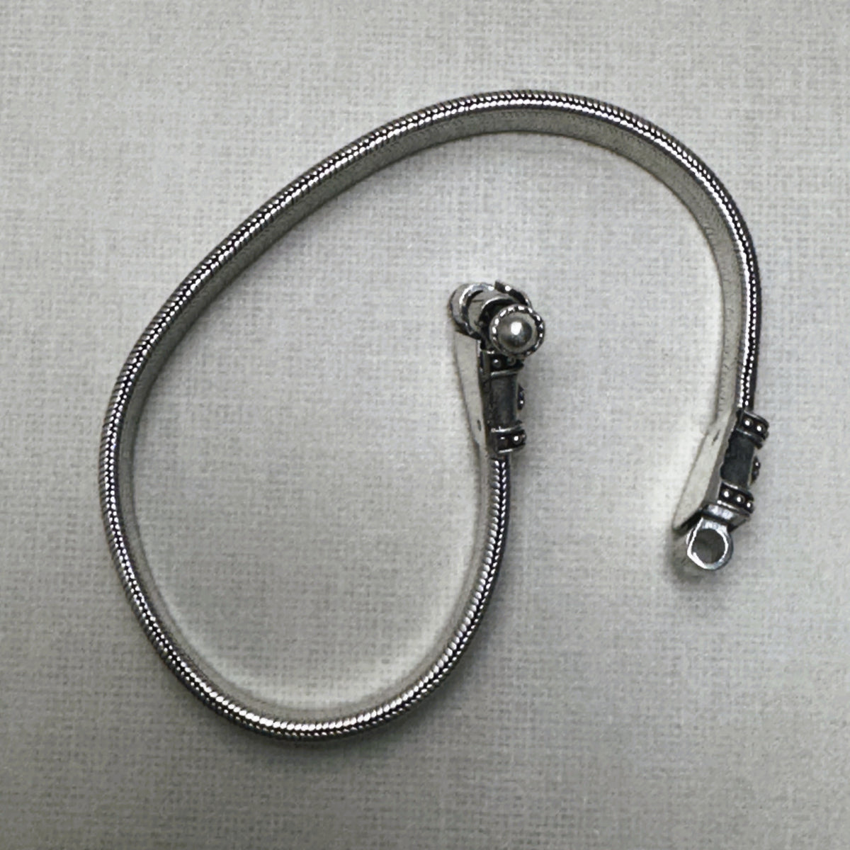 The flat snake silver anklet seen on its edge, coiled, with the clasp caps at the top.