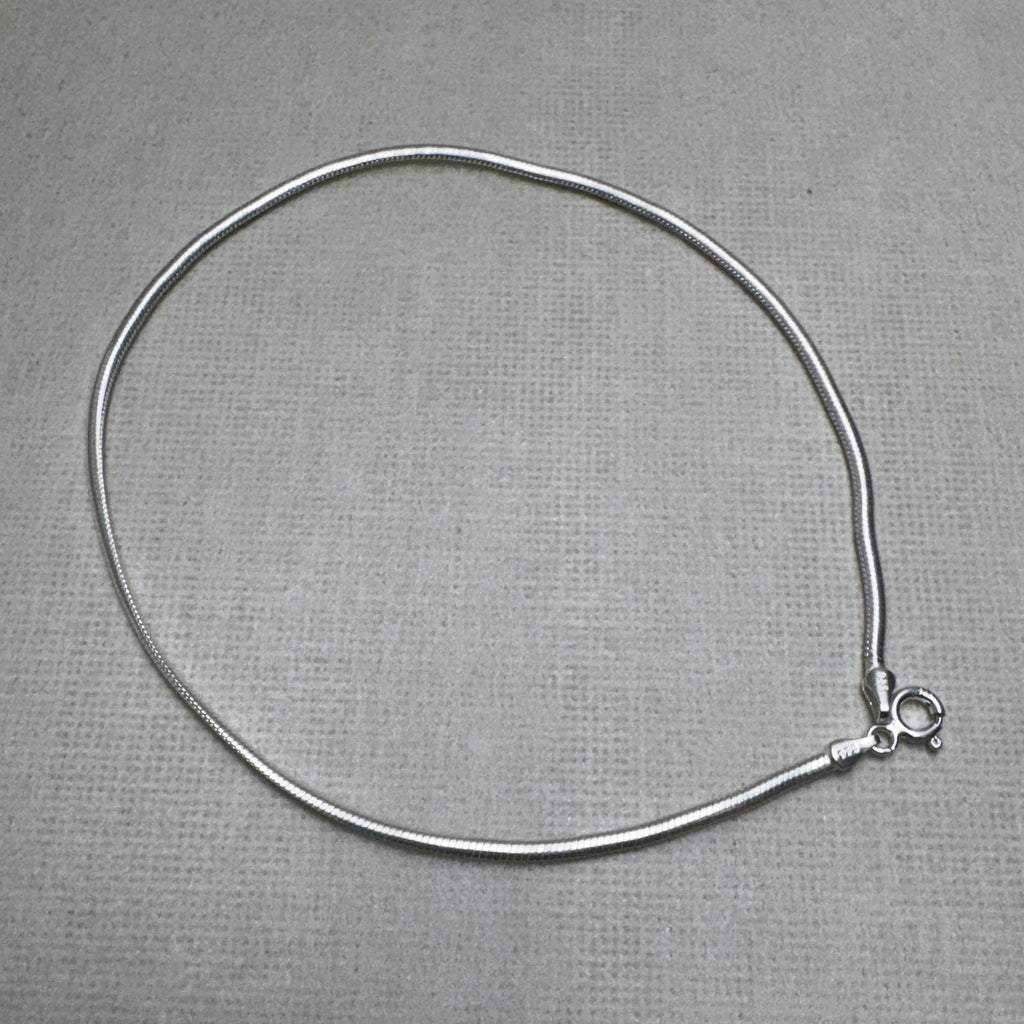 Sterling silver round snake chain anklet laid in a loop with its spring ring clasp, on a grey surface.
