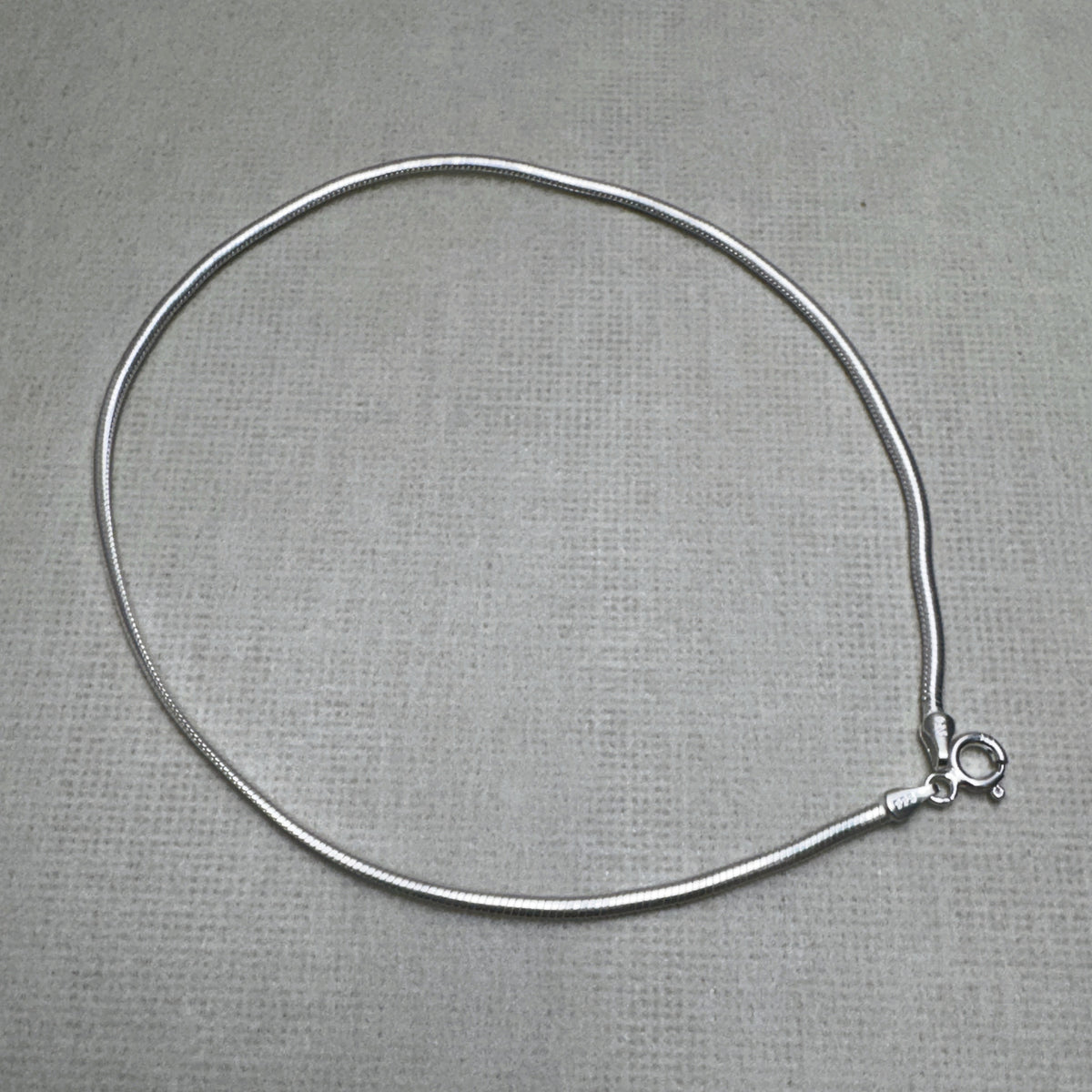Sterling silver round snake chain anklet laid in a loop with its spring ring clasp, on a grey surface.