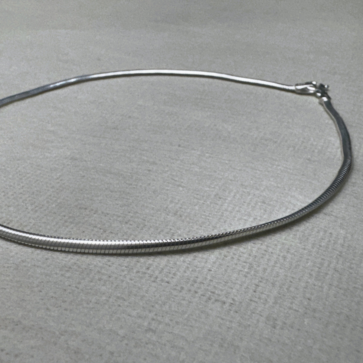 The round snake chain silver anklet laid in an oval, clasp at the right.