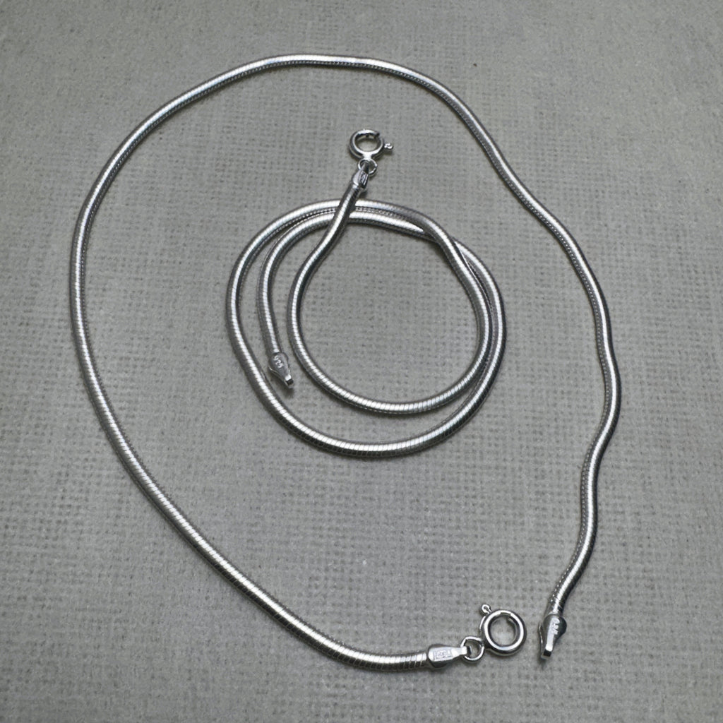 A pair of round snake chain silver anklets, one coiled inside the other.