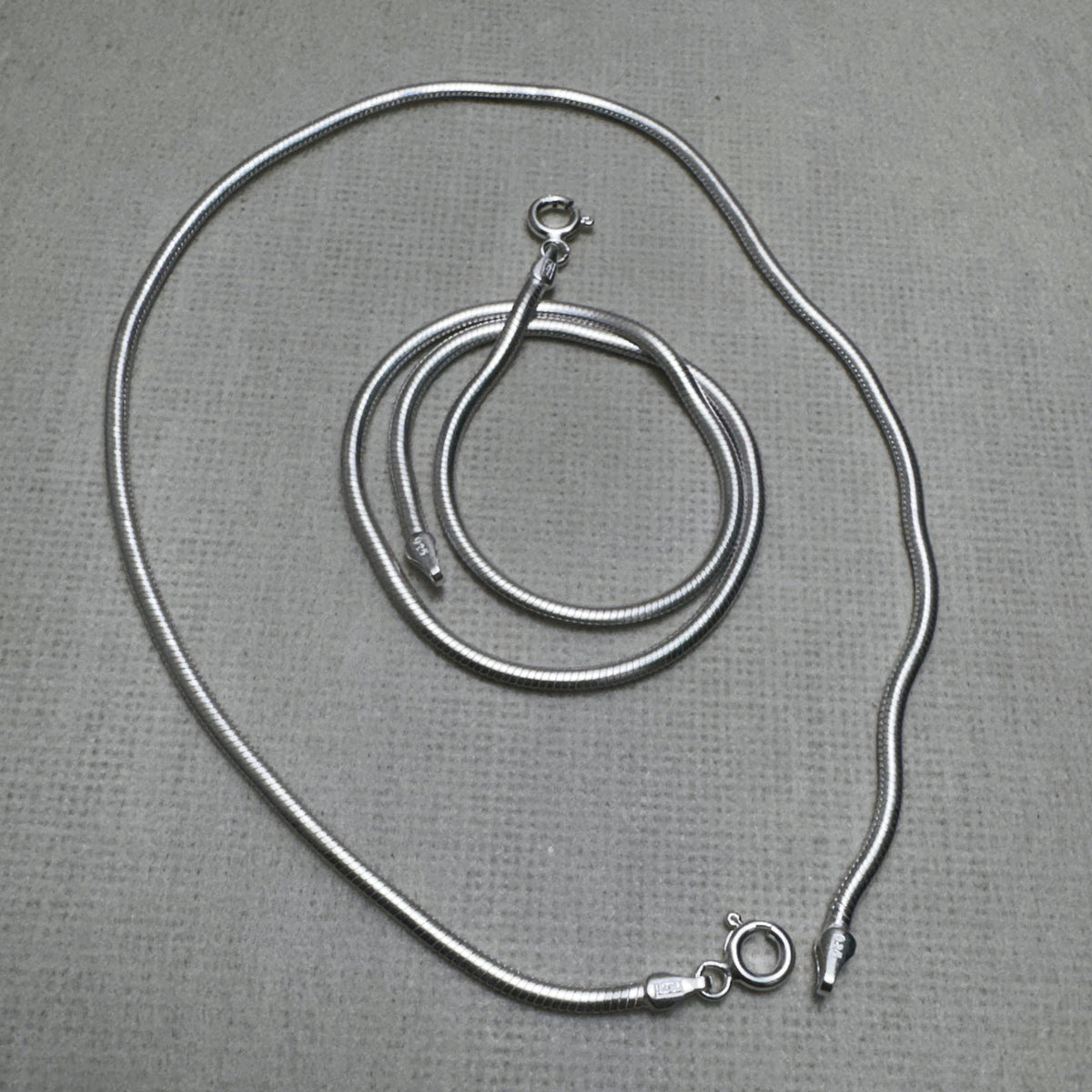 A pair of round snake chain silver anklets, one coiled inside the other.