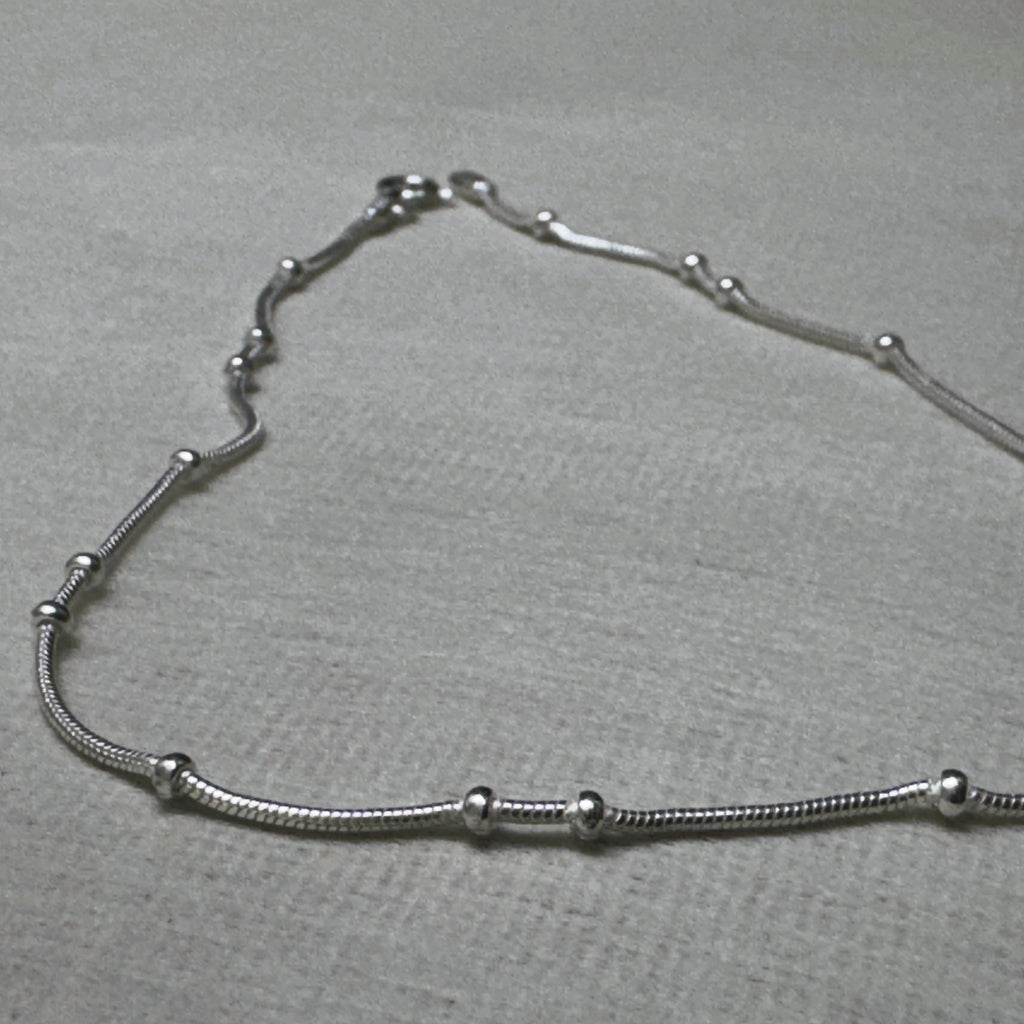 The bead station snake chain anklet laid in a curve on a grey surface.
