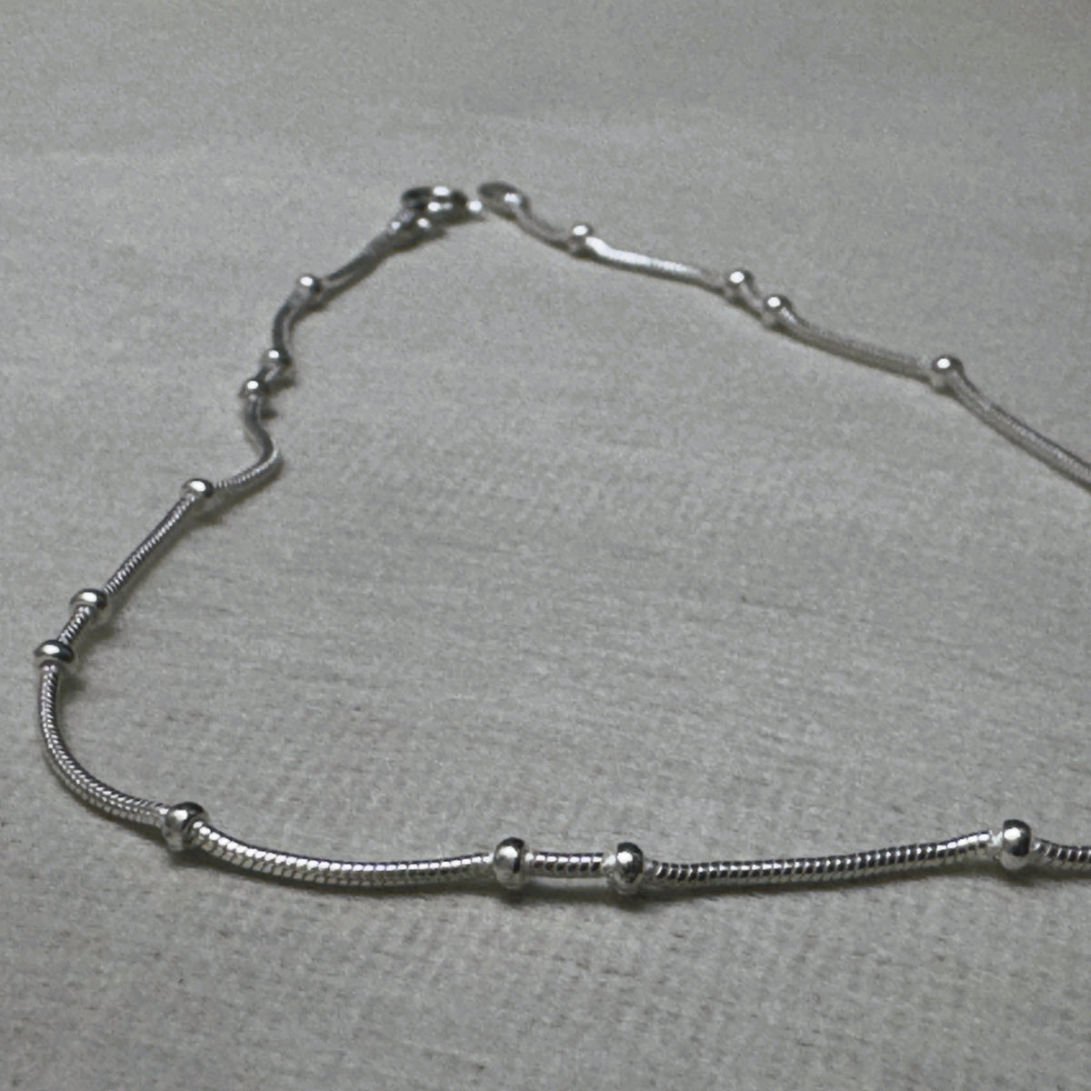The bead station snake chain anklet laid in a curve on a grey surface.
