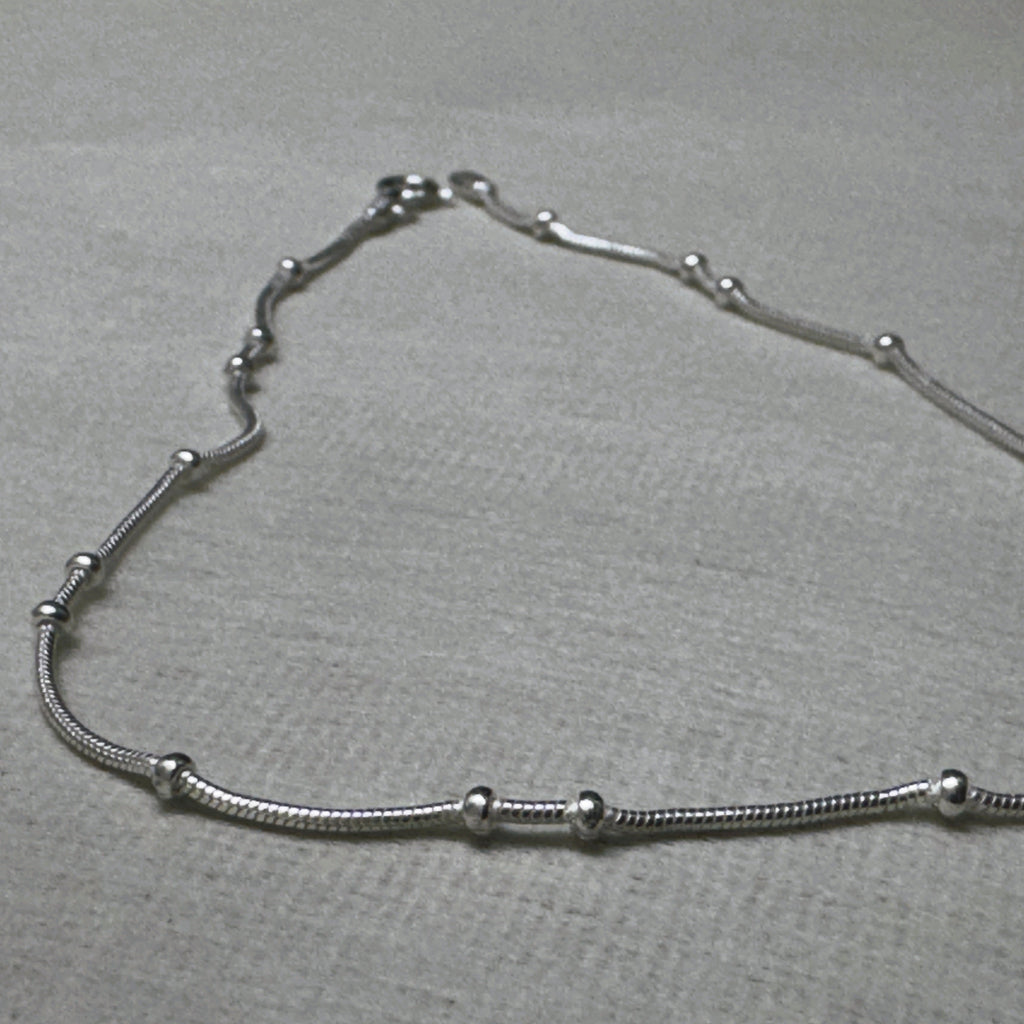 Pair of bead station snake chain silver payal: one anklet laid in a curve on a grey surface.