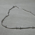 Pair of bead station snake chain silver payal: one anklet laid in a curve on a grey surface.
