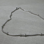 Pair of bead station snake chain silver payal: one anklet laid in a curve on a grey surface.