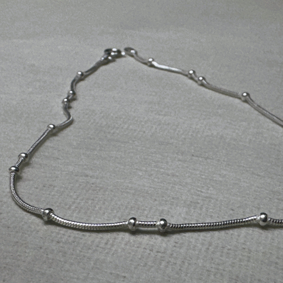 Pair of bead station snake chain silver payal: one anklet laid in a curve on a grey surface.