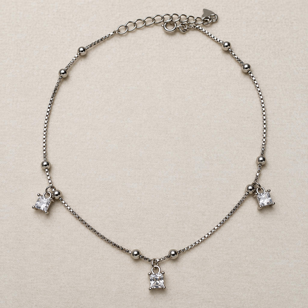 Sterling silver box chain anklet with bead stations and three princess-cut American Diamond drops, laid in a loop on a cream surface.