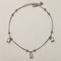 Sterling silver box chain anklet with bead stations and three princess-cut American Diamond drops, laid in a loop on a cream surface.