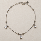 Sterling silver box chain anklet with bead stations and three princess-cut American Diamond drops, laid in a loop on a cream surface.