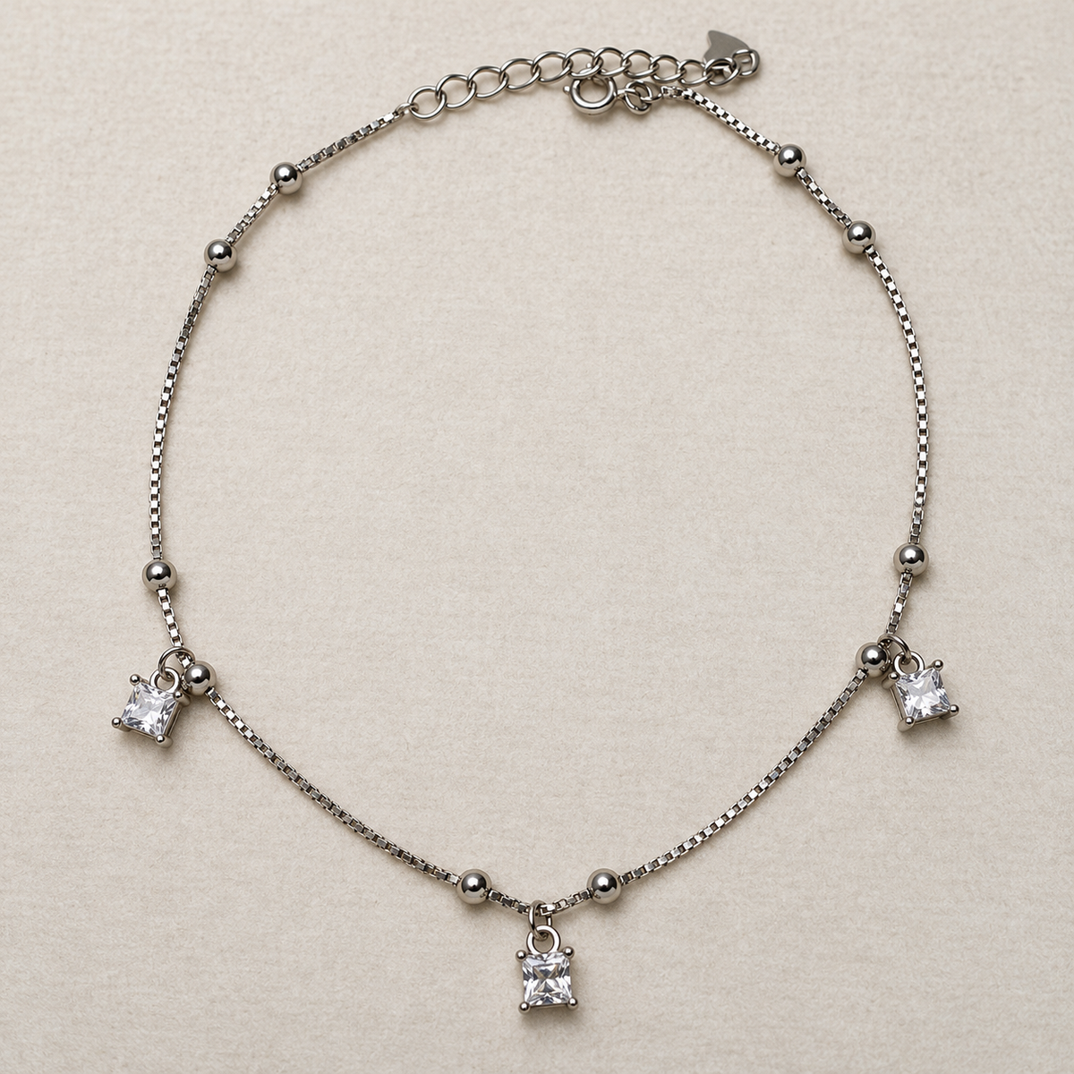 Sterling silver box chain anklet with bead stations and three princess-cut American Diamond drops, laid in a loop on a cream surface.