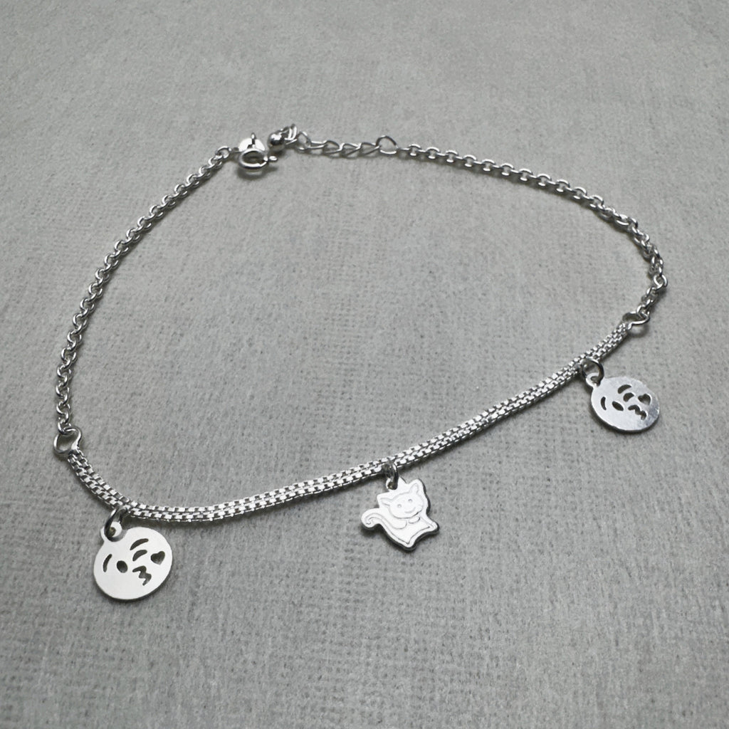 Sterling silver double rolo chain anklet with two emoji face charms and a cat charm, laid in a loop.