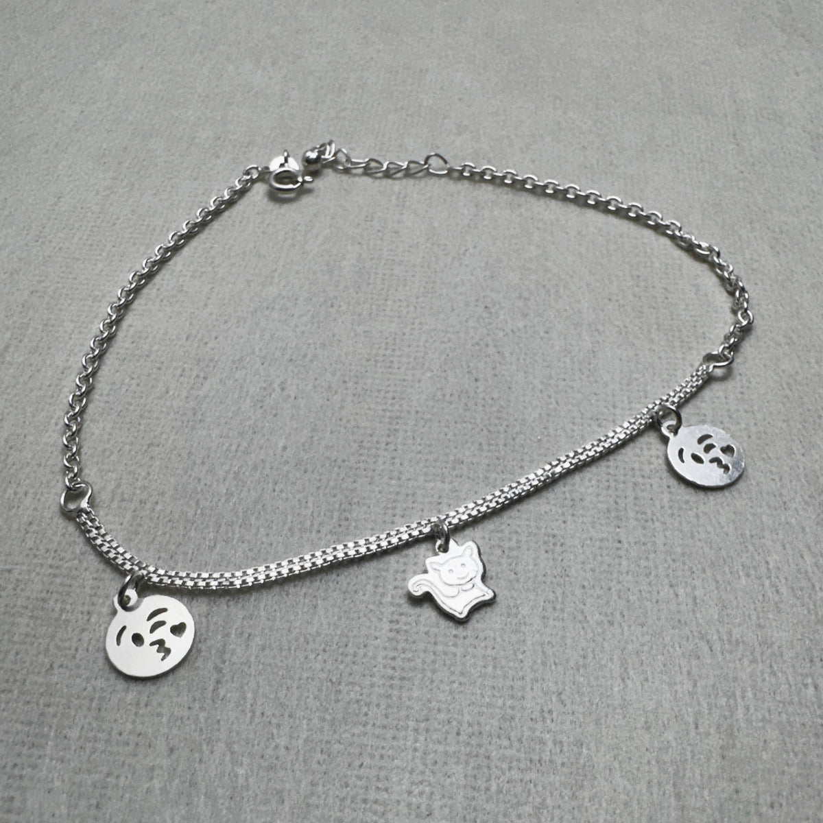 Sterling silver double rolo chain anklet with two emoji face charms and a cat charm, laid in a loop.