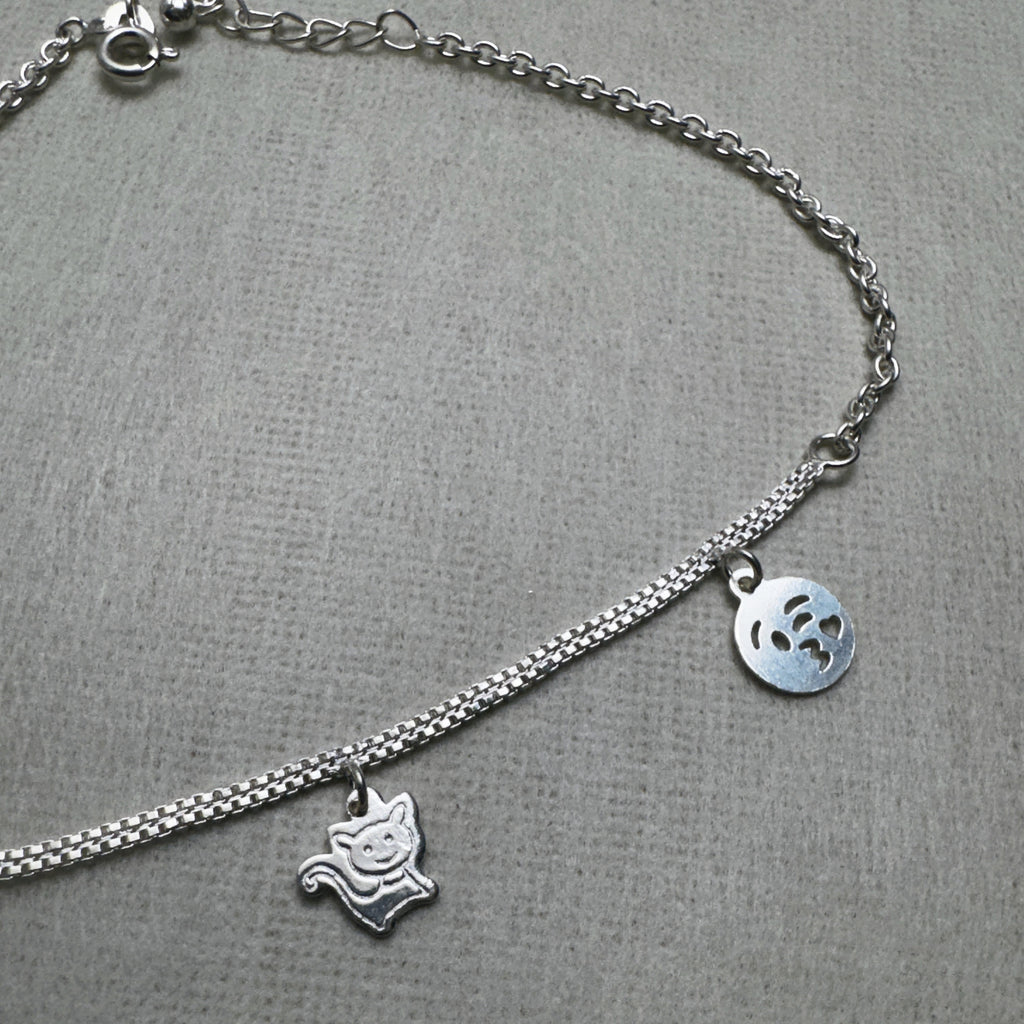 The three flat silver charms of the anklet, two emoji faces and a cat, on the double chain.