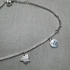 The three flat silver charms of the anklet, two emoji faces and a cat, on the double chain.