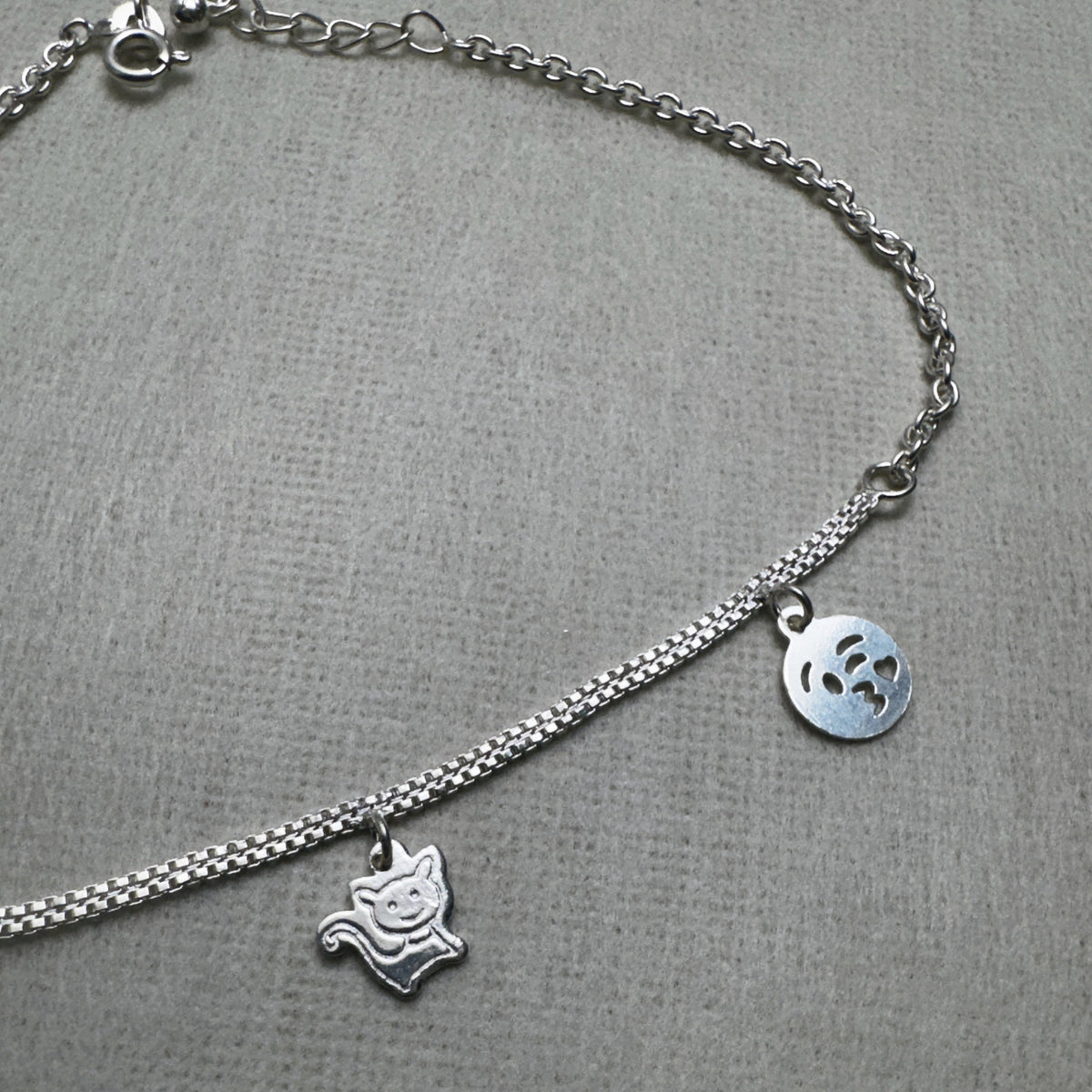 The three flat silver charms of the anklet, two emoji faces and a cat, on the double chain.