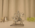 Sterling silver Durga idol seated on her tiger on a pierced base, with a silver lamp and pot behind it.