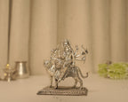 Sterling silver Durga idol seated on her tiger on a pierced base, with a silver lamp and pot behind it.