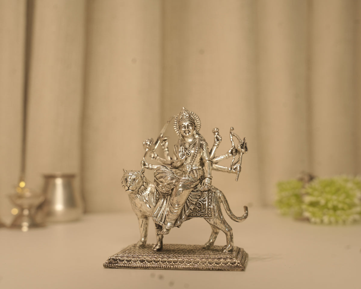 Sterling silver Durga idol seated on her tiger on a pierced base, with a silver lamp and pot behind it.