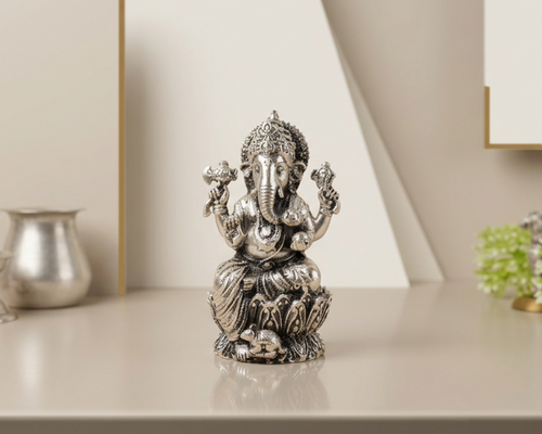 Antique finish sterling silver Ganesh idol on a lotus base, on a pale table with a silver pot and a mirror behind.